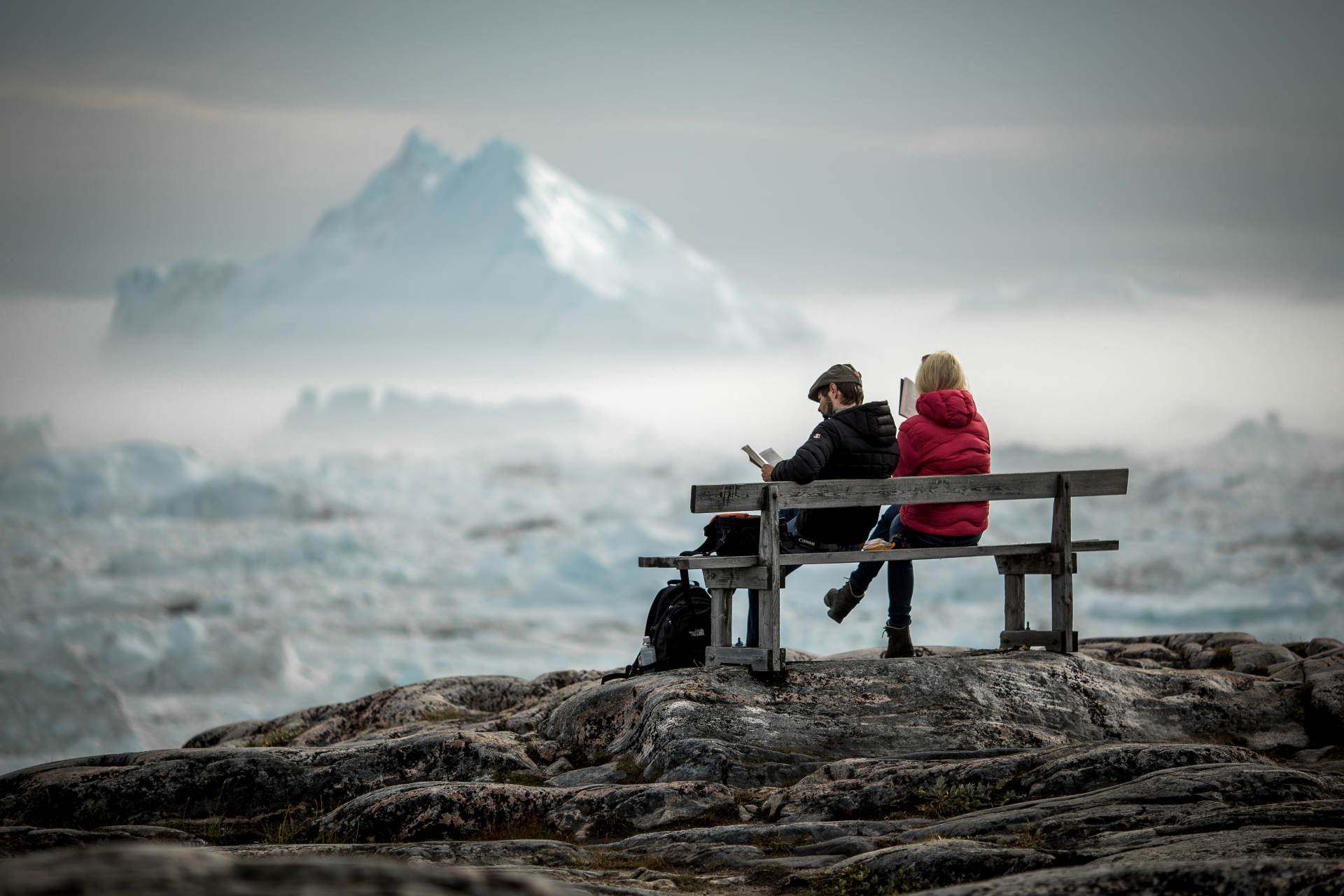 Two Travelers Reading On A Bench Overlooking Icebergs In The Ilulissat Ice Fjord In Greenland